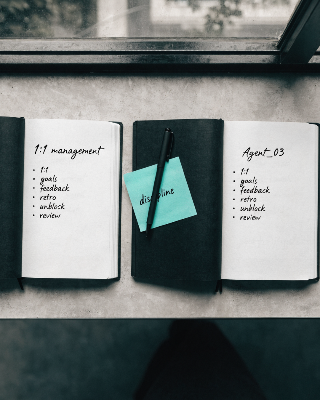 Two open notebooks side by side: one titled '1:1 management', the other 'Agent_03'. Both list the same bullets — 1:1, goals, feedback, retro, unblock, review. Between them, a sticky note reading 'discipline' with a pen on top.